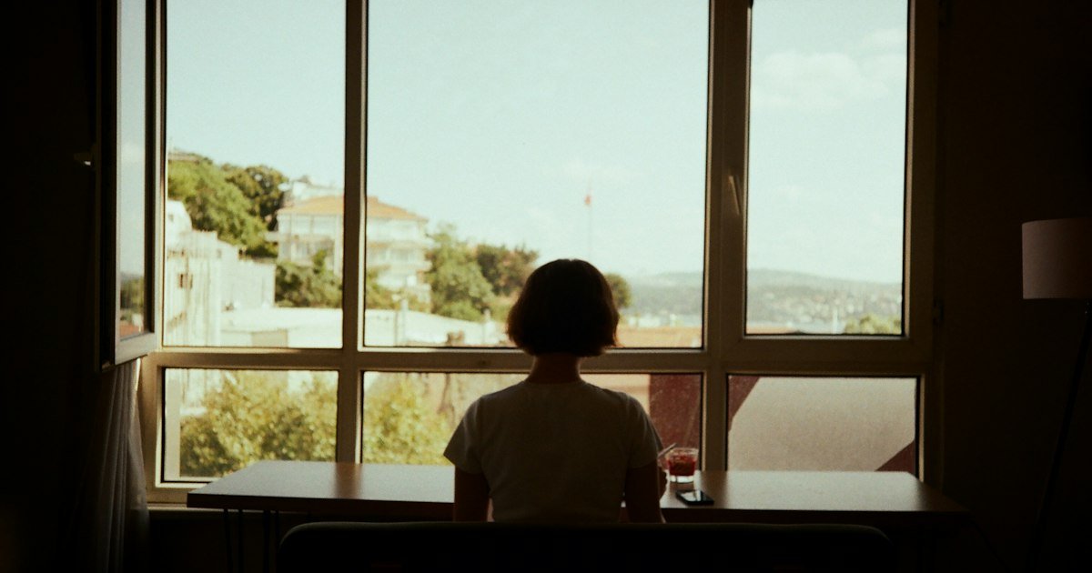 Person sitting alone at a table, looking out of a window, lost in thought